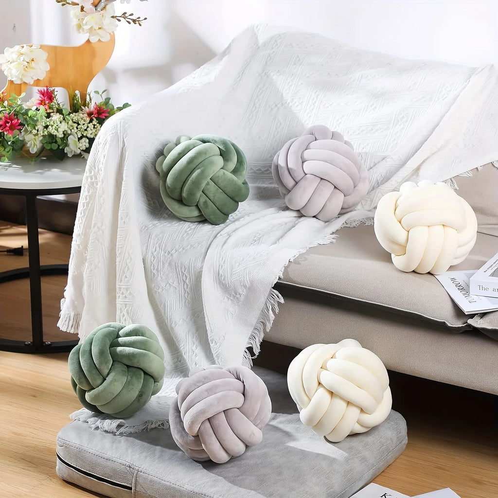 Decorative knot pillows in green, gray, and cream on a sofa with a blanket and flowers in the background.