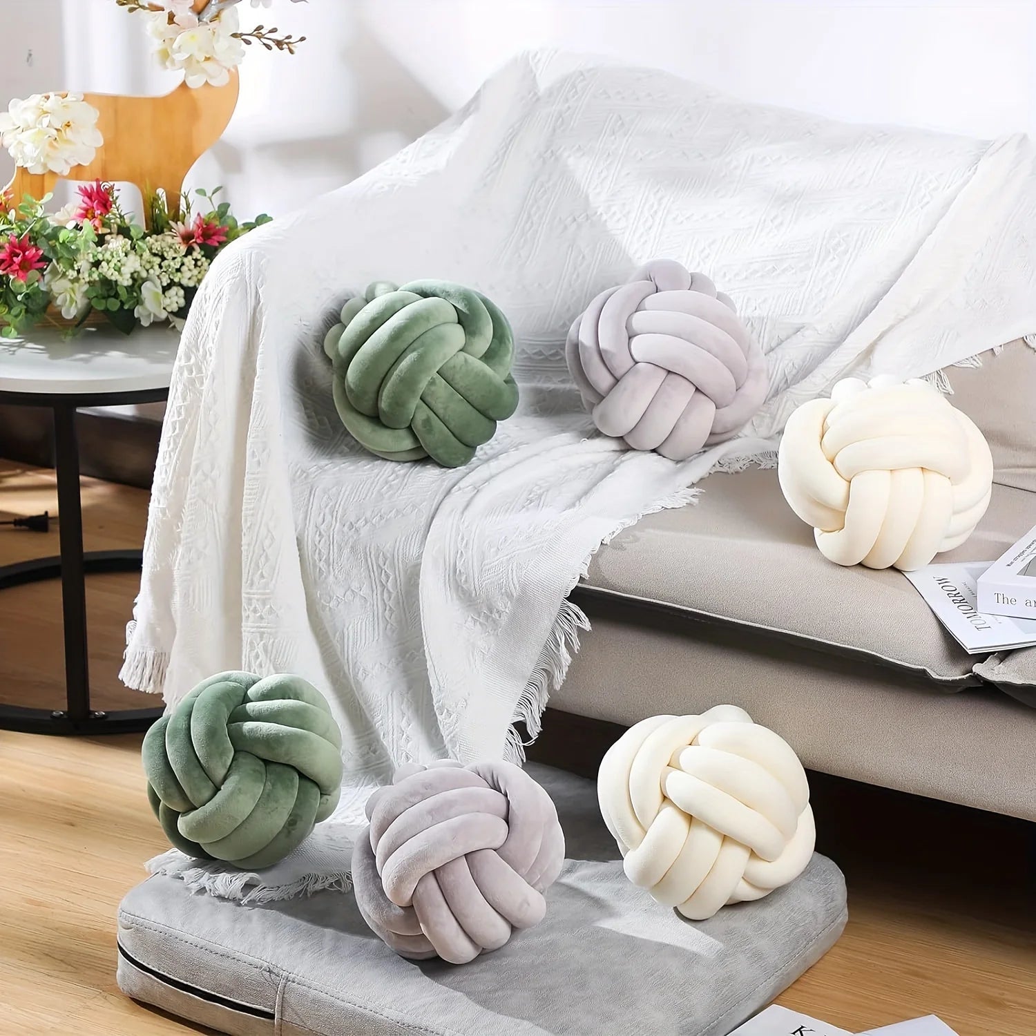 Decorative knot pillows in green, gray, and cream on a sofa with a blanket and flowers in the background.