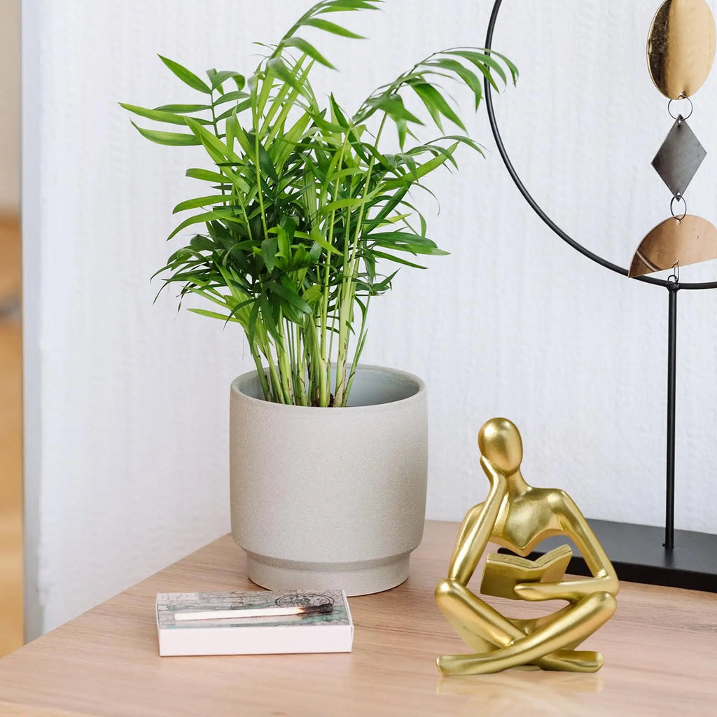 Gold Thinker Reading Sculpture sitting with a book on a beige background of a person sitting on a wooden surface with a potted plant and decorative lamp in the background.