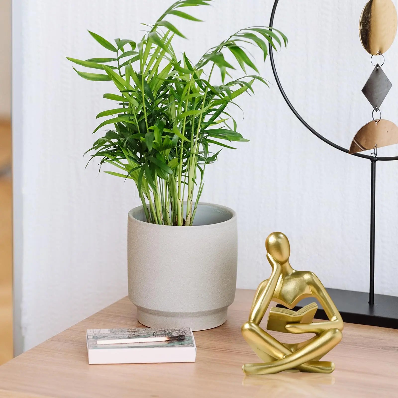 Gold Thinker Reading Sculpture sitting with a book on a beige background of a person sitting on a wooden surface with a potted plant and decorative lamp in the background.