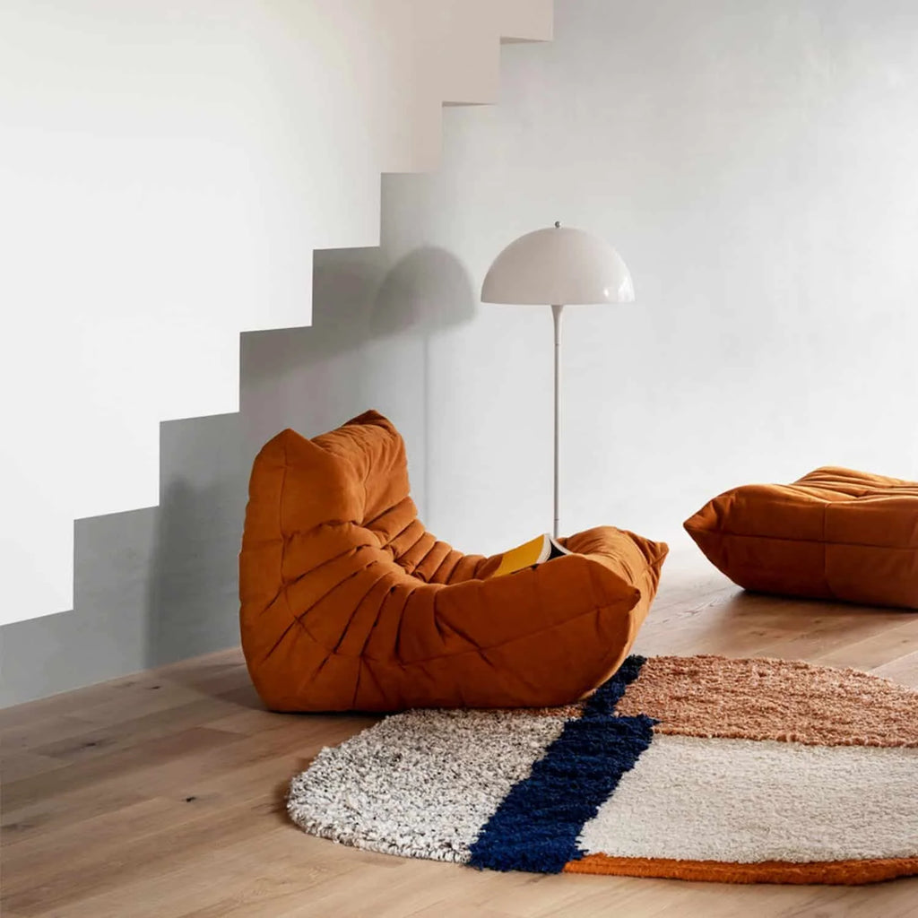 One seater Orange Corduroy Caterpillar Lazy Sofa with a cushion on a wooden floor with a lamp and colorful rug in the background.