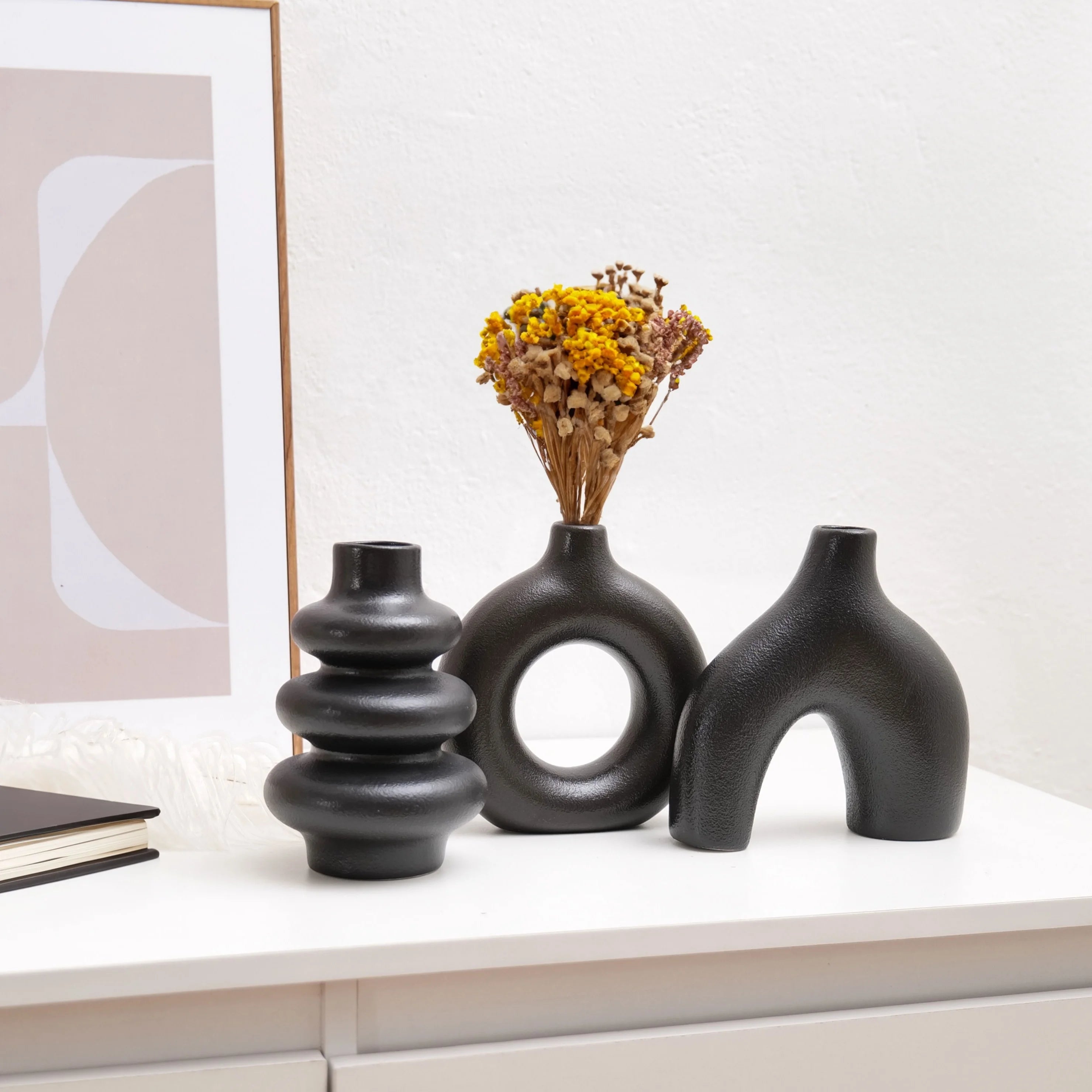 Three black Boho Ceramic Vases on a white surface with a framed abstract art piece in the background.