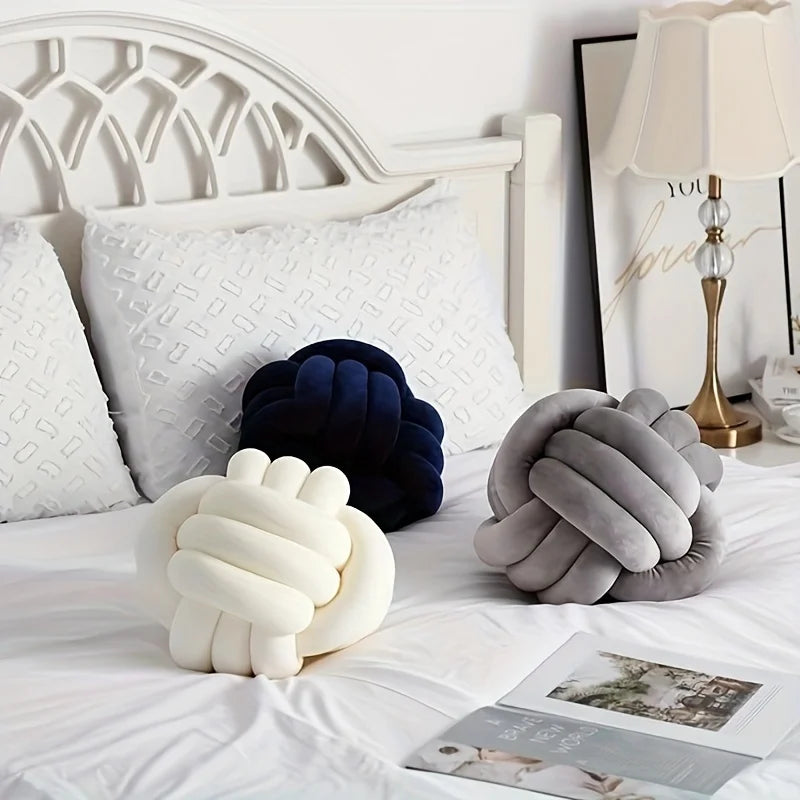 Three knot-shaped decorative pillows in white, navy, and gray on a bed with a lamp in the background.