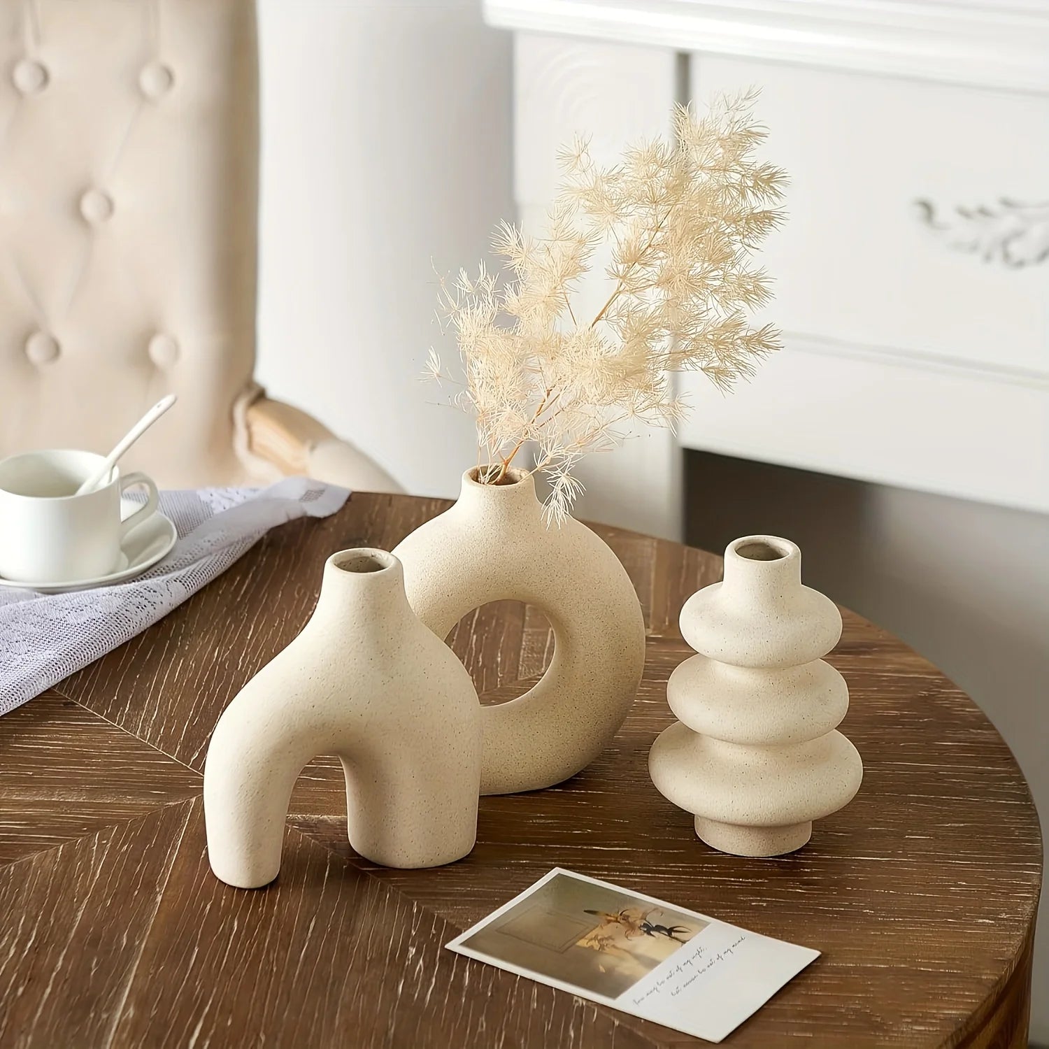Three light yellow Boho Ceramic Vases on a wooden table with a card and a cup in the background.